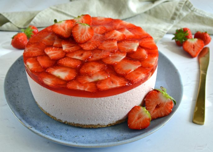 Opskrift cheesecake med jordbær på en blå tallerken på et bord med en guldkniv og et beige viskestykke