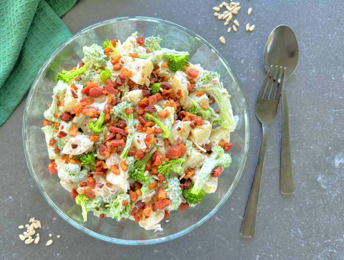 Opskrift broccoli kartoffelsalat med bacontern på toppen, anrettet i en glasskål med sort bestik