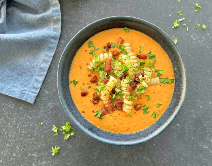 Opskrift nem rodfrugtsuppe anrettet med bacontern, suppehorn og et drys persille i en blå skål på et sort bord